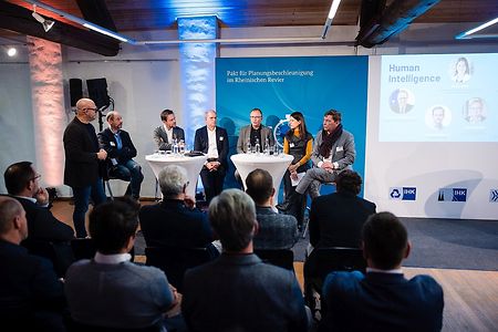 Sieben Personen diskutieren auf einer B&uuml;hne, davor Publikum.